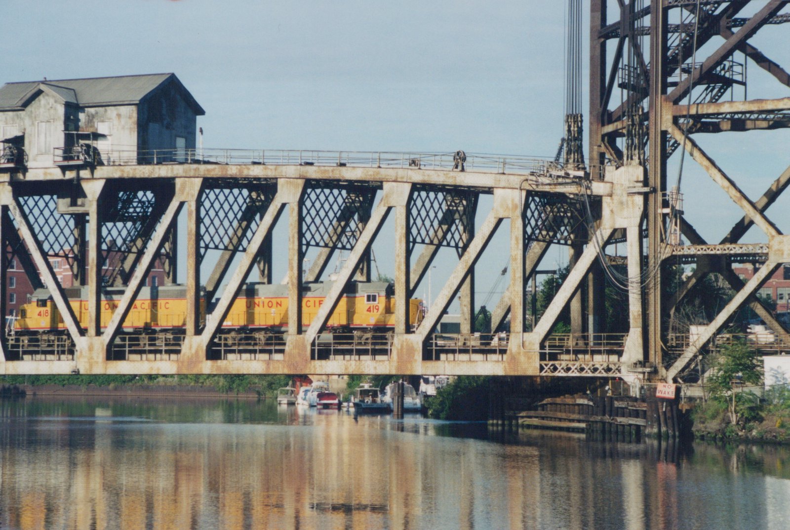 Two yellow and gray Union Pacific E9 locomotives numbered 418 and 419 travel across a steel truss bridge over a river, with buildings in the background and boats moored below.