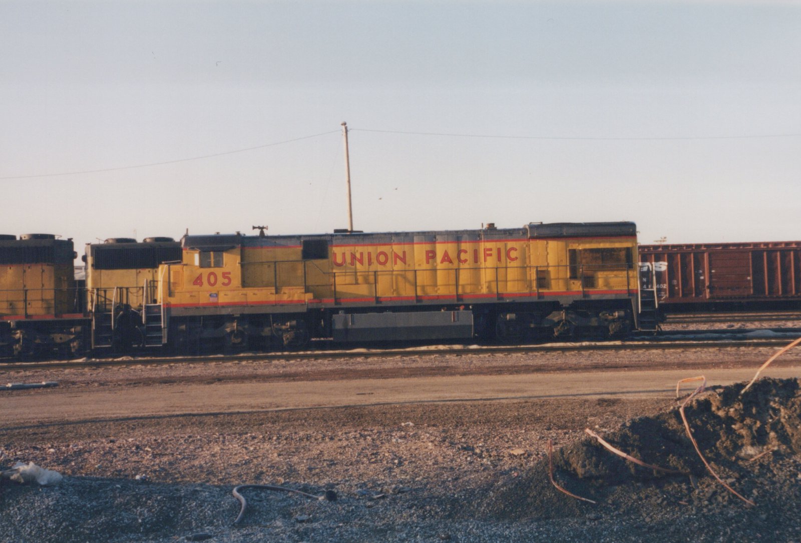 Side view of a yellow Union Pacific GP9 diesel locomotive, number 405, on a track in a gravel yard with a brown Norfolk Southern freight car visible behind it.