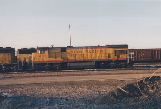 Side view of a yellow Union Pacific GP9 diesel locomotive, number 405, on a track in a gravel yard with a brown Norfolk Southern freight car visible behind it.