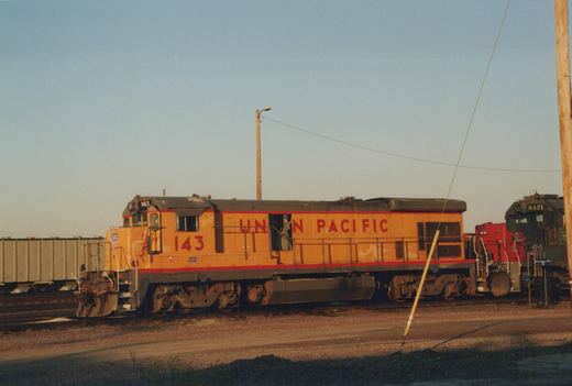 A yellow Union Pacific diesel locomotive numbered 143 on a railroad track in a yard, with another locomotive numbered 8321 visible behind it.