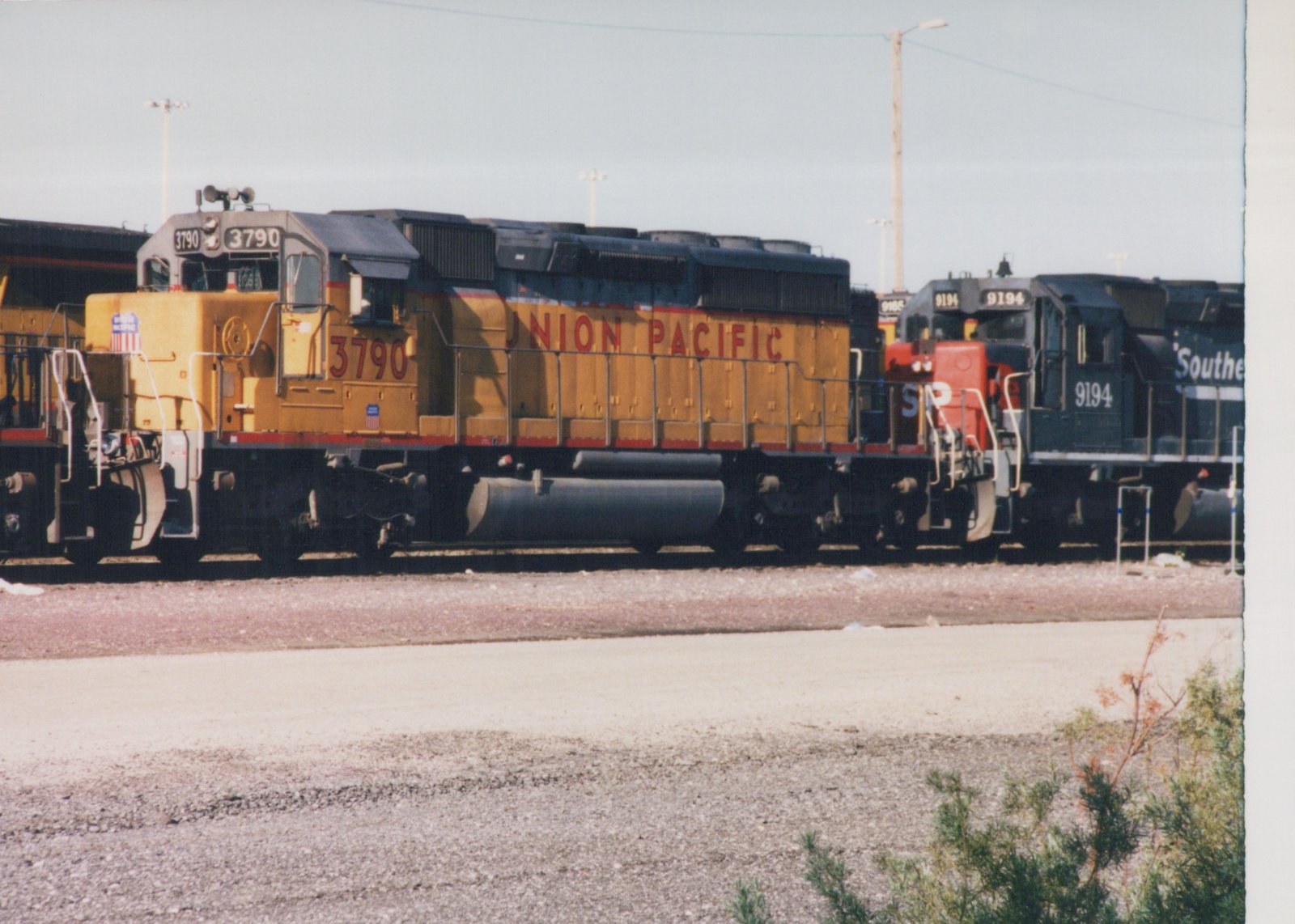 Side view of two diesel locomotives, a yellow Union Pacific 3790 and a black Southern Pacific 9194, parked side by side on a railroad track in a gravel yard under a clear sky.