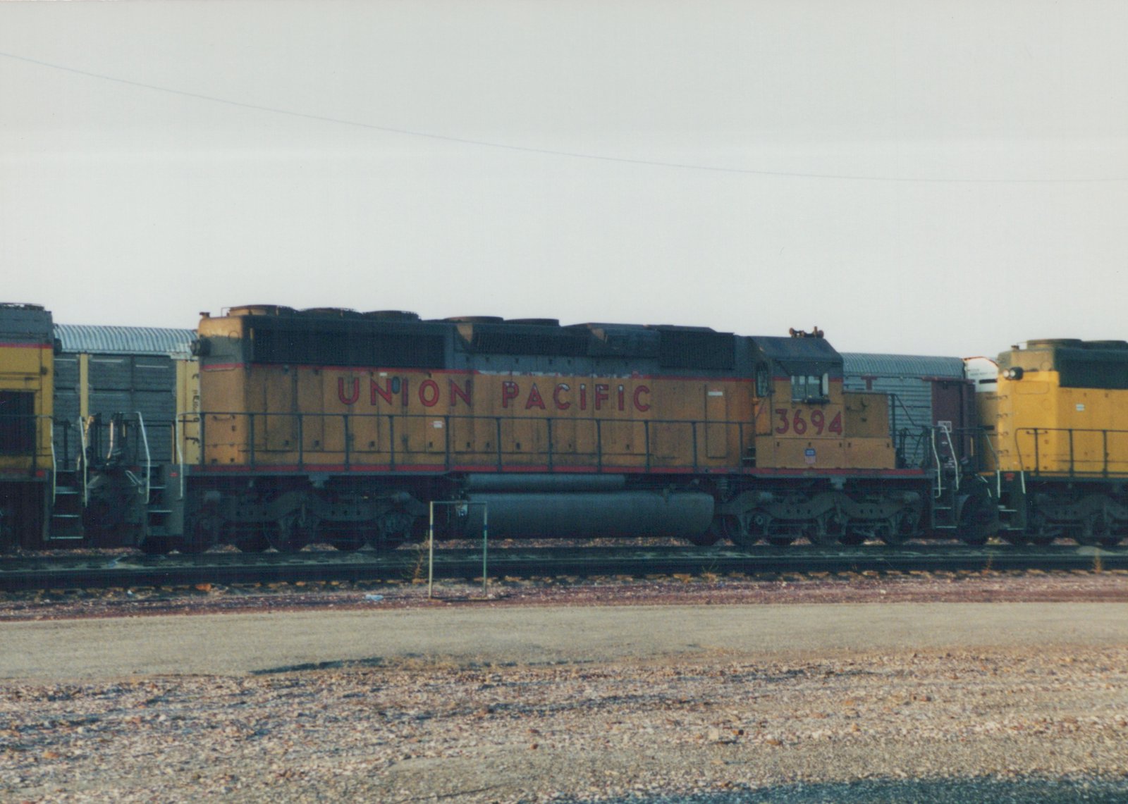 Side view of a Union Pacific GP38-2 diesel locomotive, number 3694, in a gravel yard with freight cars behind it, under an overcast sky.