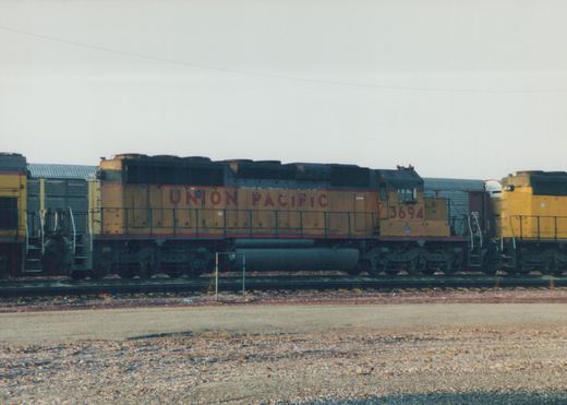 Side view of a Union Pacific GP38-2 diesel locomotive, number 3694, in a gravel yard with freight cars behind it, under an overcast sky.