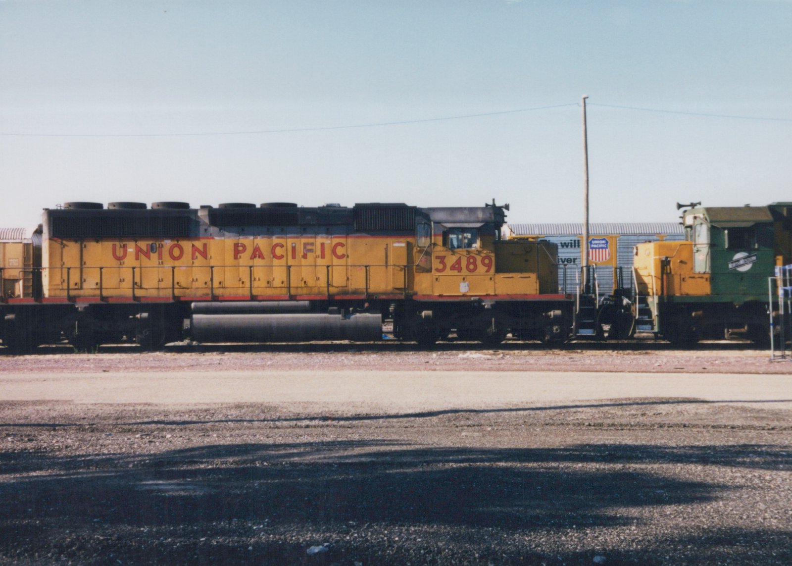 A yellow Union Pacific diesel locomotive numbered 3489 sits on tracks in a gravel yard with a green and yellow switcher and a blue Union Pacific boxcar in the background.