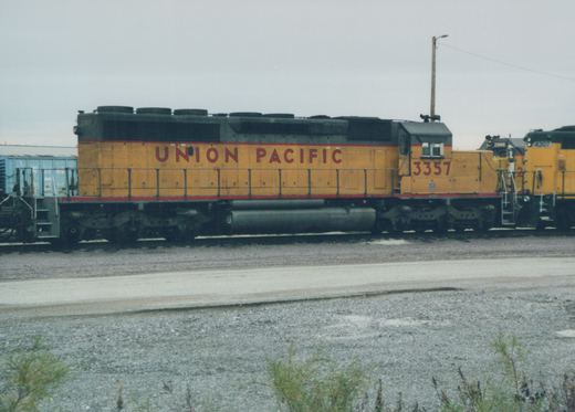 Side view of a Union Pacific locomotive number 3357 in a gravel yard with other locomotives and a blue freight car in the background under an overcast sky.