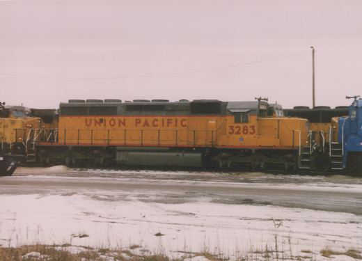 A yellow Union Pacific diesel locomotive with the number 3283 is parked in a snowy yard alongside other locomotives under an overcast sky.
