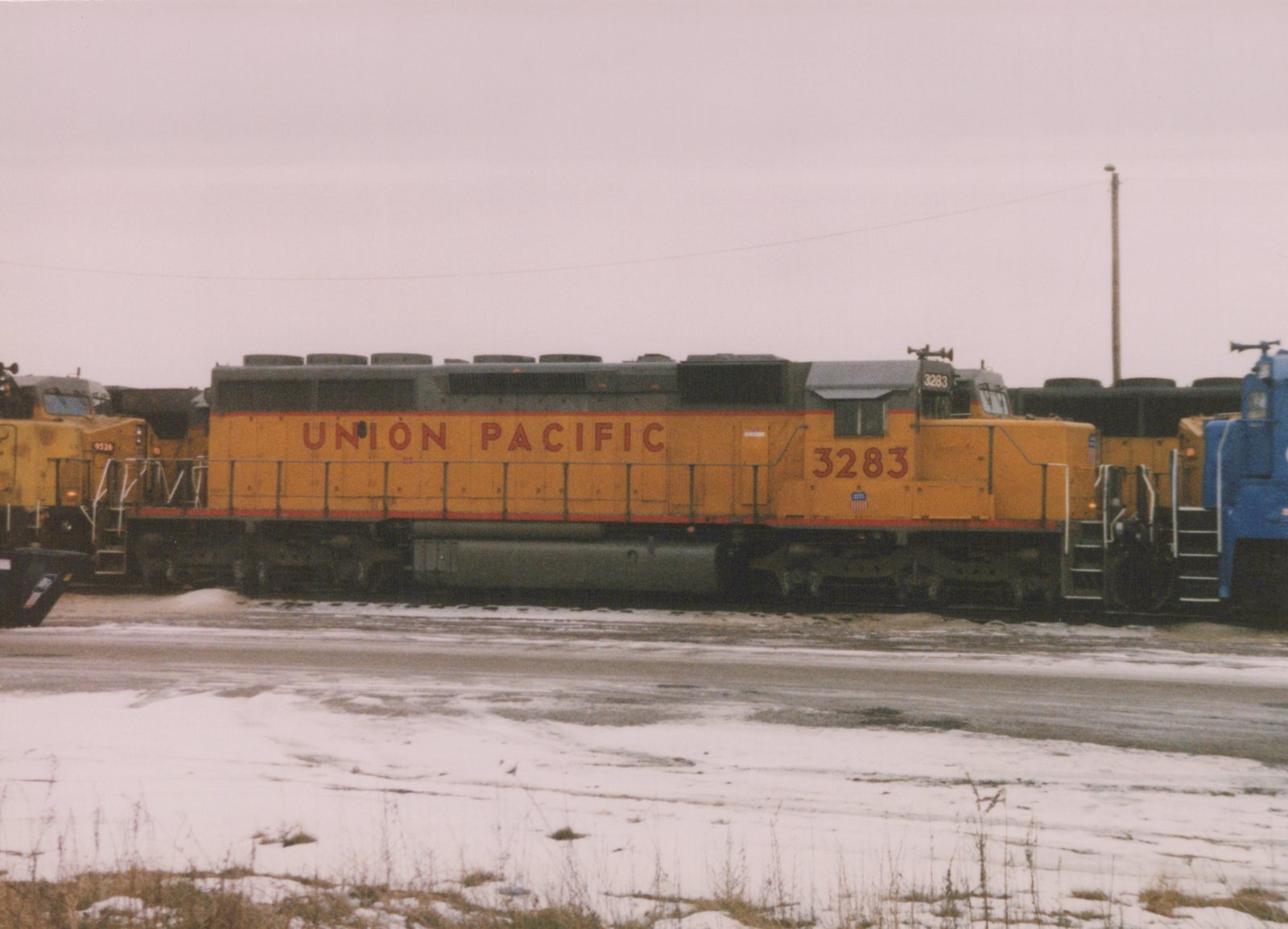 A yellow Union Pacific diesel locomotive with the number 3283 is parked in a snowy yard alongside other locomotives under an overcast sky.