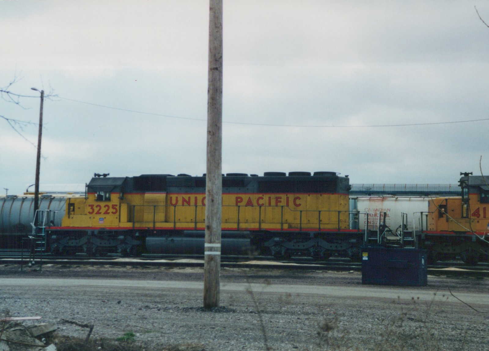 A side view of a yellow and black Union Pacific diesel locomotive, number 3225, parked on tracks in a rail yard next to other railcars on an overcast day.