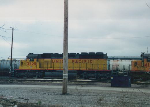 A side view of a yellow and black Union Pacific diesel locomotive, number 3225, parked on tracks in a rail yard next to other railcars on an overcast day.