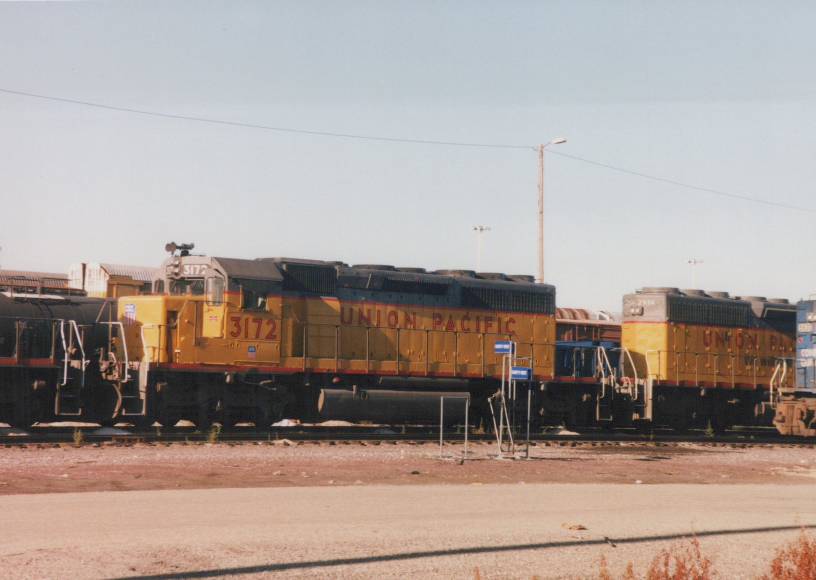 Two yellow Union Pacific diesel locomotives, numbered 3172 and 2974, are parked side-by-side in a gravel yard with other freight cars in the background under a clear blue sky.