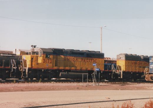 Two yellow Union Pacific diesel locomotives, numbered 3172 and 2974, are parked side-by-side in a gravel yard with other freight cars in the background under a clear blue sky.