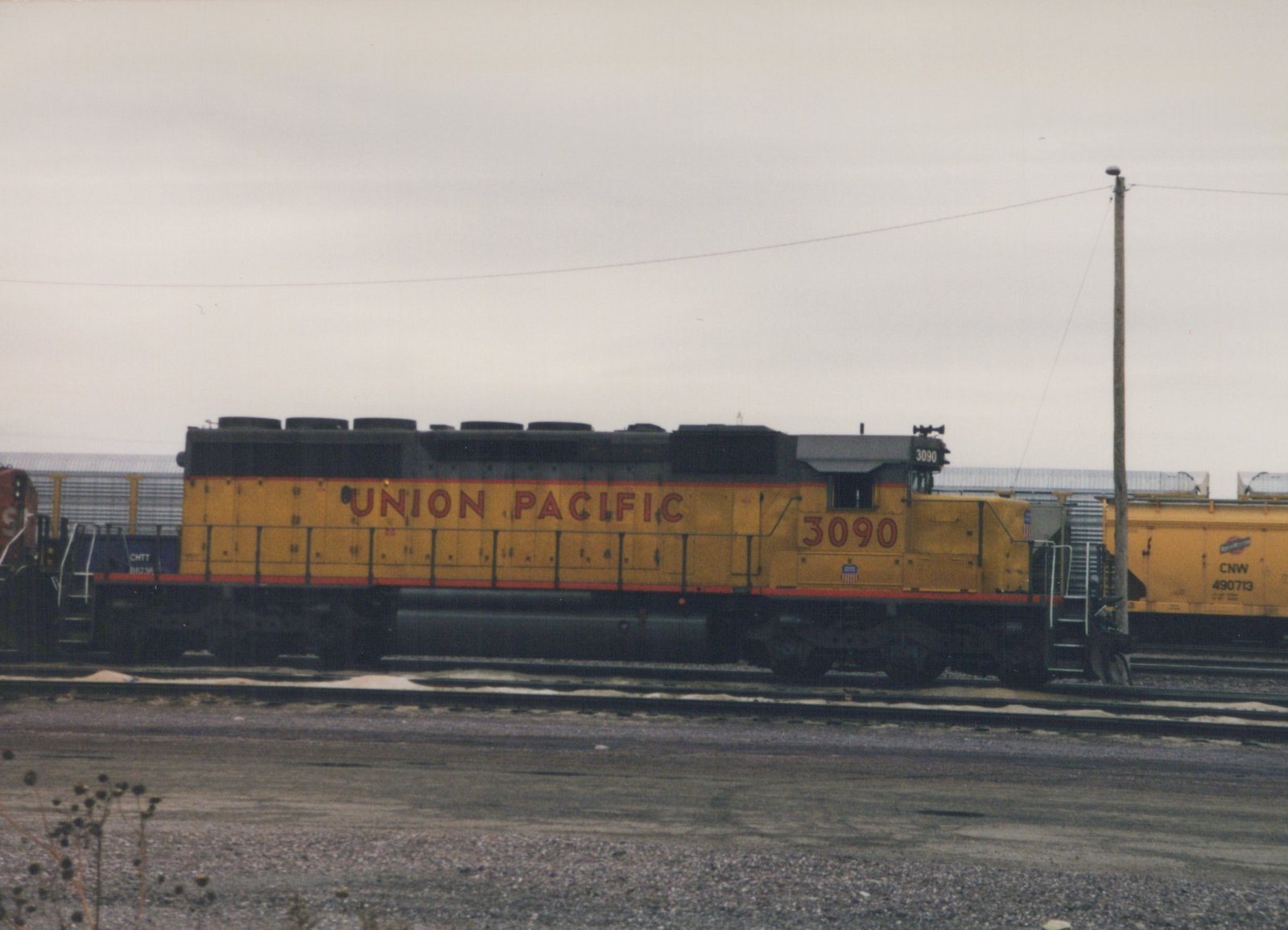 A yellow and black Union Pacific locomotive numbered 3090 is parked on tracks in a yard, with other freight cars visible behind it and a cloudy sky above.