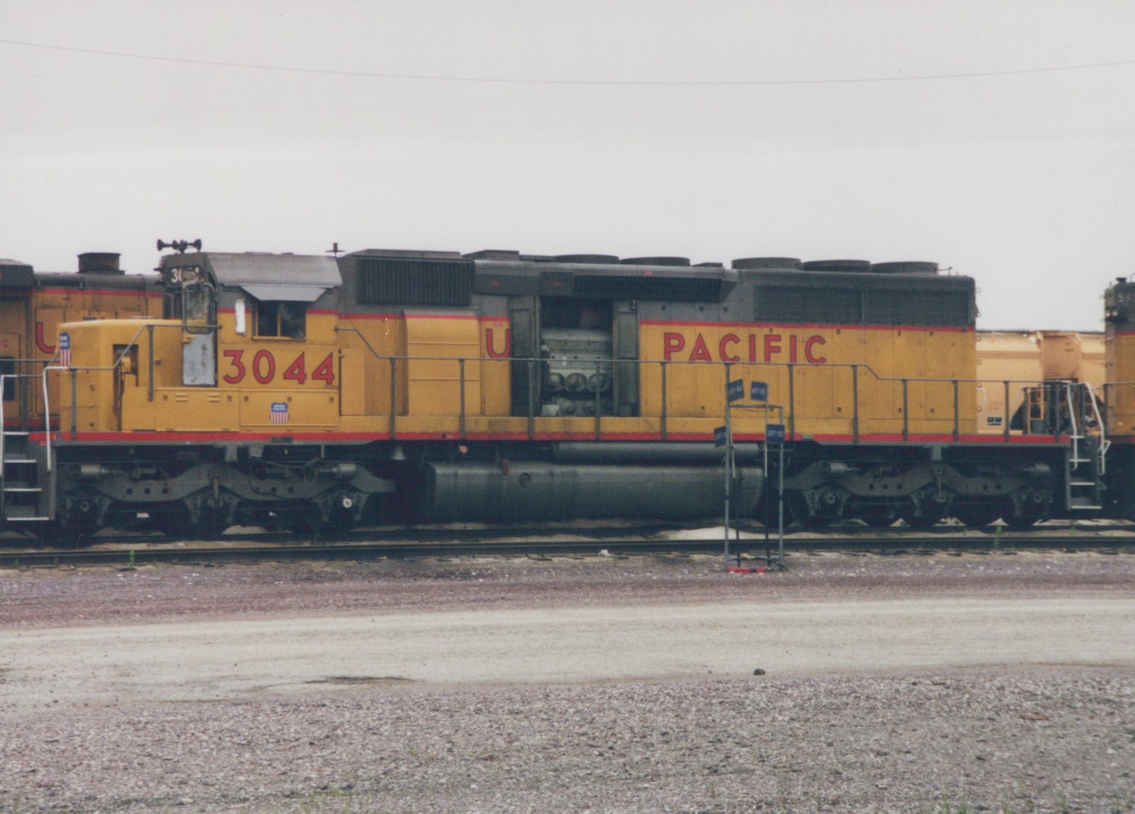 A yellow and red Union Pacific diesel locomotive with the number 3044 is positioned on tracks in a gravel yard under an overcast sky.