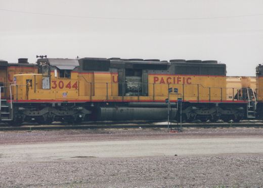 A yellow and red Union Pacific diesel locomotive with the number 3044 is positioned on tracks in a gravel yard under an overcast sky.
