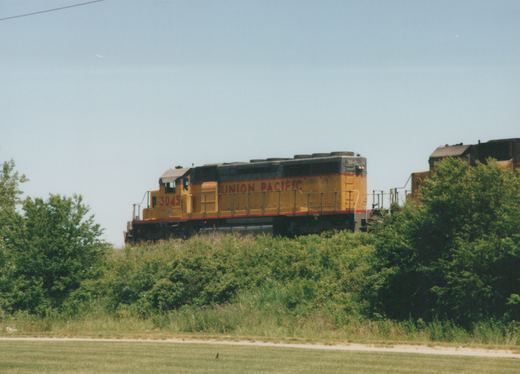A yellow and gray Union Pacific locomotive with the number 3045 on its side is seen traveling through a grassy area with trees in the background.