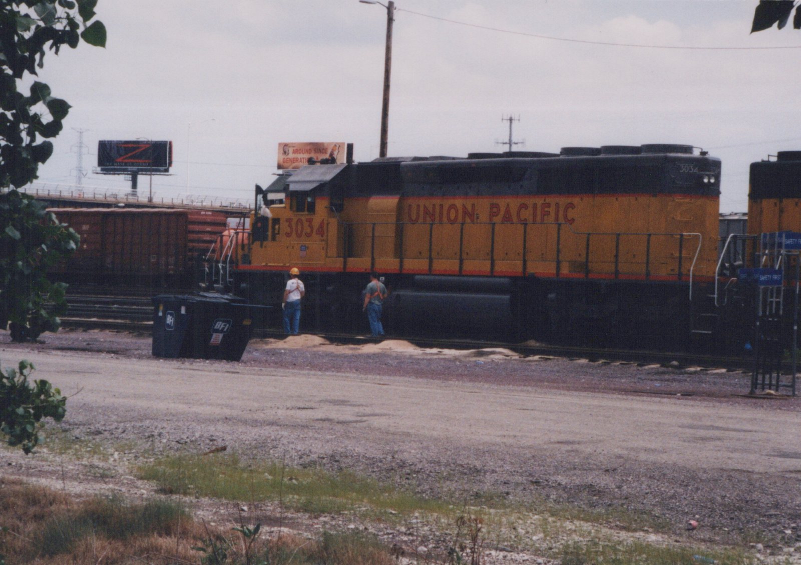 A yellow and black Union Pacific locomotive numbered 3034 sits on tracks in a gravel yard under an overcast sky, with two workers nearby and freight cars in the background.