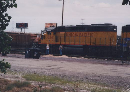 A yellow and black Union Pacific locomotive numbered 3034 sits on tracks in a gravel yard under an overcast sky, with two workers nearby and freight cars in the background.