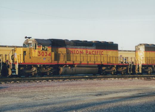 Side view of a yellow Union Pacific locomotive with the number 3034 and red lettering, on a railroad track in a gravel yard with snow on the ground.