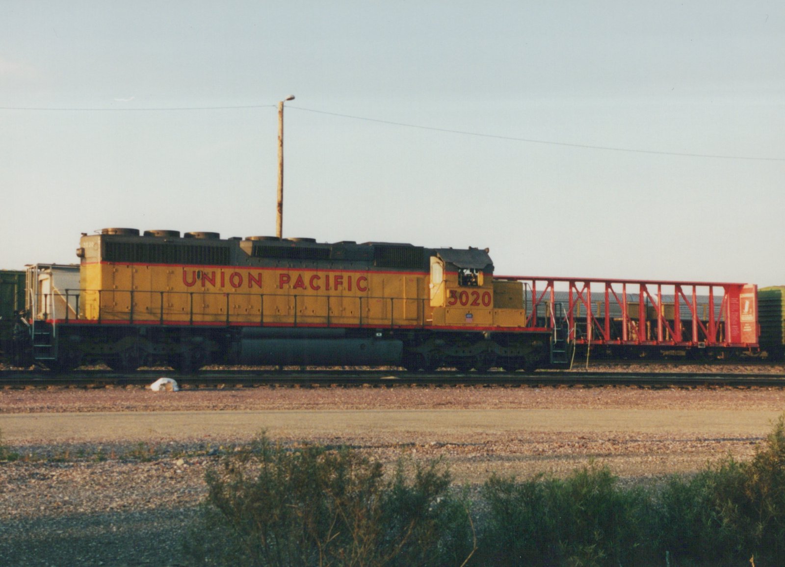 A yellow and red Union Pacific diesel locomotive, number 3020, sits on tracks next to a red freight car, viewed from the side in a sunny yard.