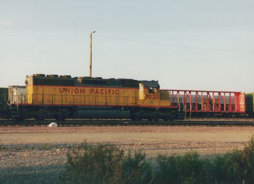 A yellow and red Union Pacific diesel locomotive, number 3020, sits on tracks next to a red freight car, viewed from the side in a sunny yard.
