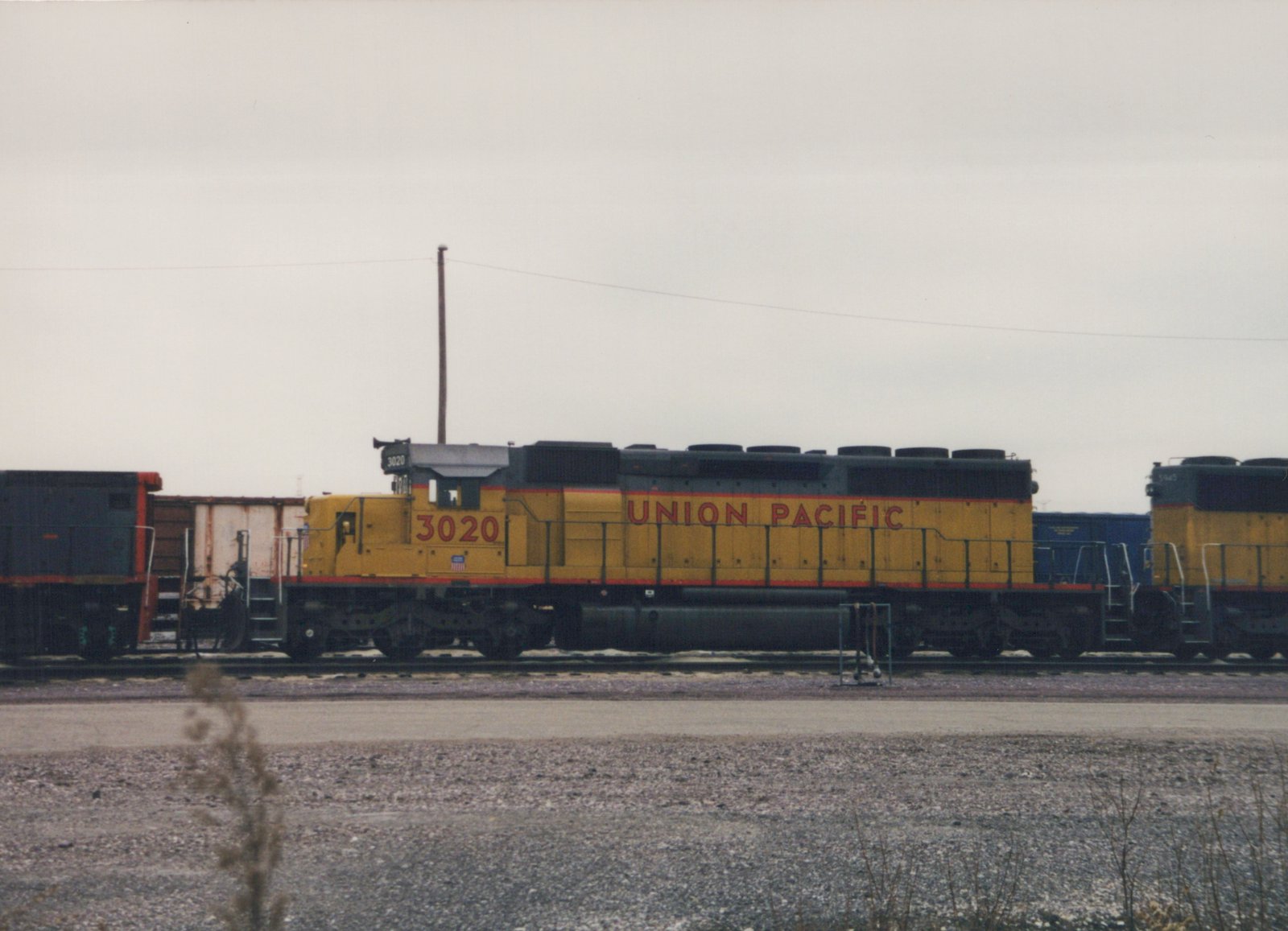 A yellow and black Union Pacific diesel locomotive numbered 3020 is parked on a track in a rail yard with patches of snow on the ground and another locomotive visible behind it.