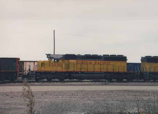 A yellow and black Union Pacific diesel locomotive numbered 3020 is parked on a track in a rail yard with patches of snow on the ground and another locomotive visible behind it.