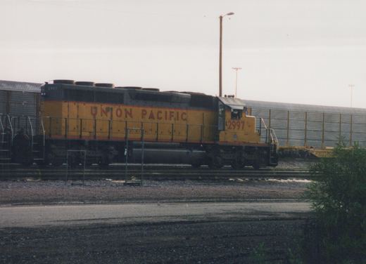 A yellow and gray Union Pacific diesel locomotive numbered 2997 sits on tracks in a gravel yard, with a freight car behind it under an overcast sky.
