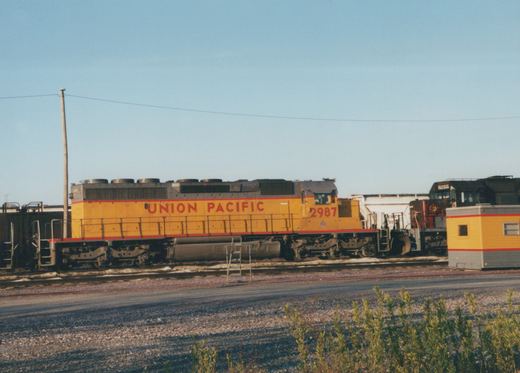 A Union Pacific diesel locomotive, number 2987, painted yellow with red stripes, is parked in a rail yard with another locomotive in the background.