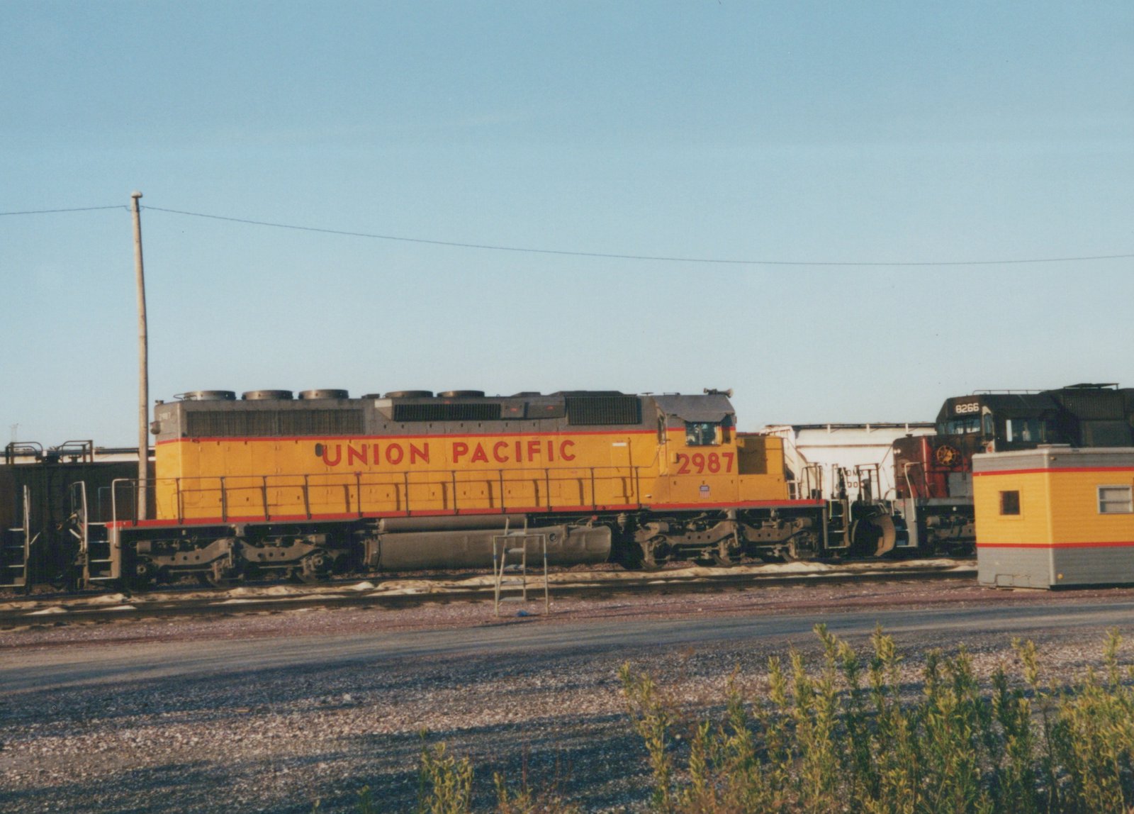A Union Pacific diesel locomotive, number 2987, painted yellow with red stripes, is parked in a rail yard with another locomotive in the background.