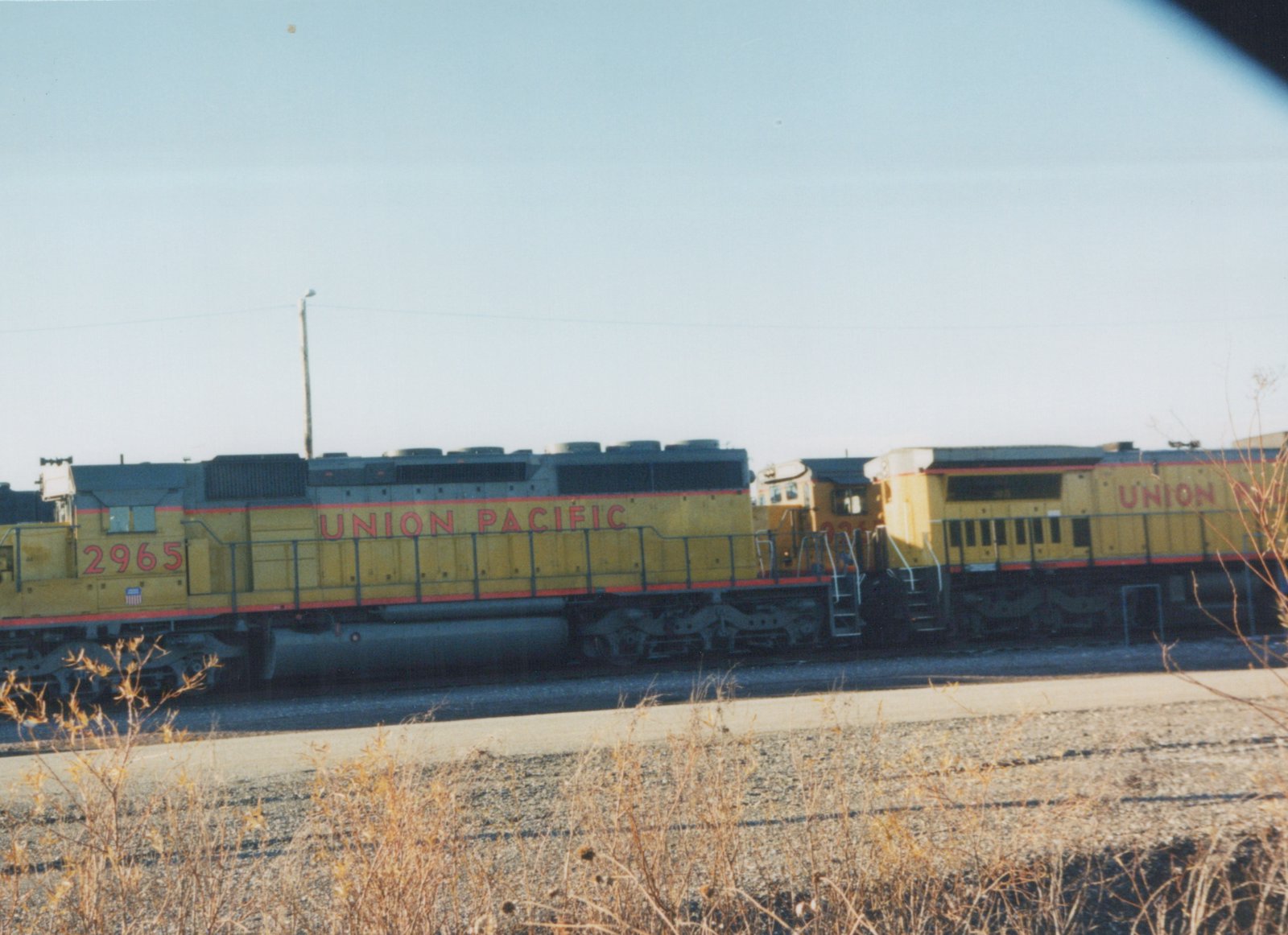 Side view of a yellow Union Pacific diesel locomotive, number 2965, with another locomotive behind it, parked on railroad tracks with dry brush in the foreground.