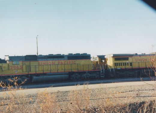 Side view of a yellow Union Pacific diesel locomotive, number 2965, with another locomotive behind it, parked on railroad tracks with dry brush in the foreground.
