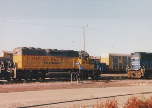 Two diesel locomotives, one yellow Union Pacific and one blue Conrail, stand on parallel tracks in a rail yard on a sunny day.