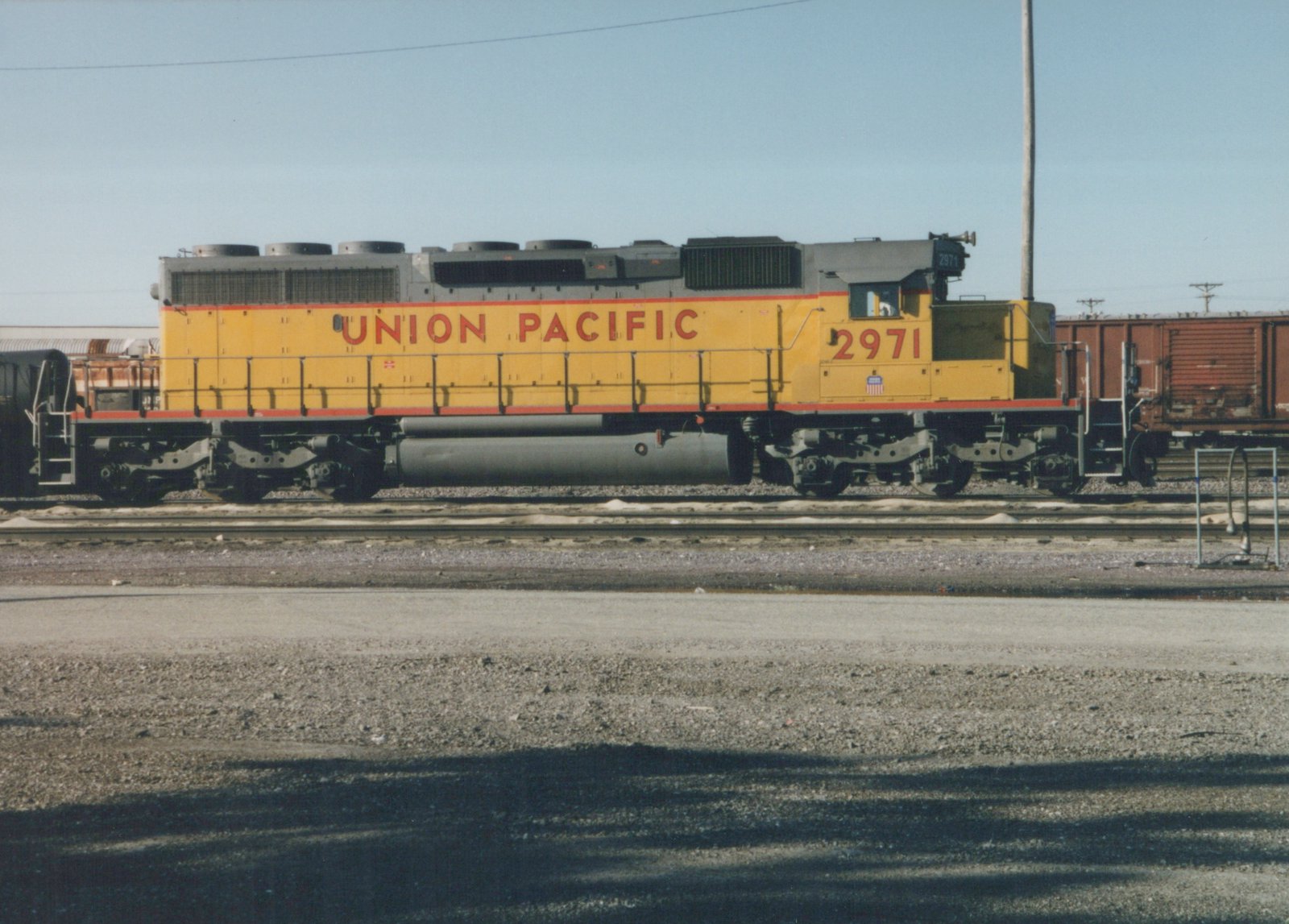 A side view of a yellow and gray Union Pacific diesel locomotive numbered 2971, sitting on tracks in a gravel yard with freight cars nearby.