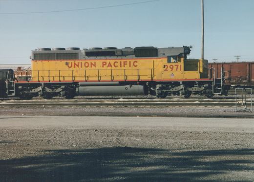 A side view of a yellow and gray Union Pacific diesel locomotive numbered 2971, sitting on tracks in a gravel yard with freight cars nearby.