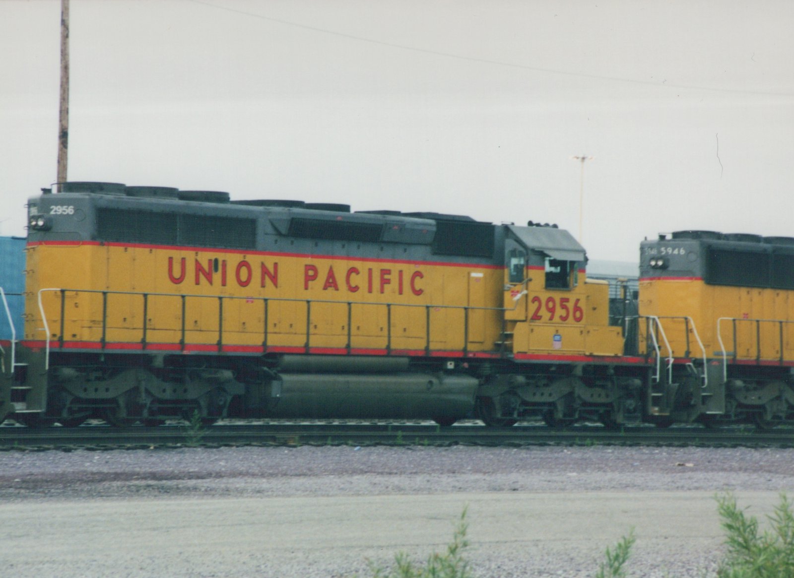Side view of two yellow and gray Union Pacific diesel locomotives, numbers 2956 and 5946, parked on parallel tracks in a gravel yard under a cloudy sky.