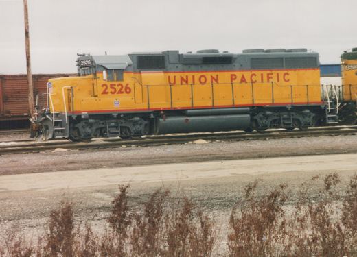 Side view of a yellow and gray Union Pacific diesel locomotive, number 2526, on a set of tracks in a yard with bare winter brush in the foreground.