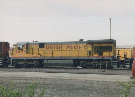 Side view of a yellow and gray Union Pacific locomotive number 2499 parked in a gravel yard with other equipment visible in the background.