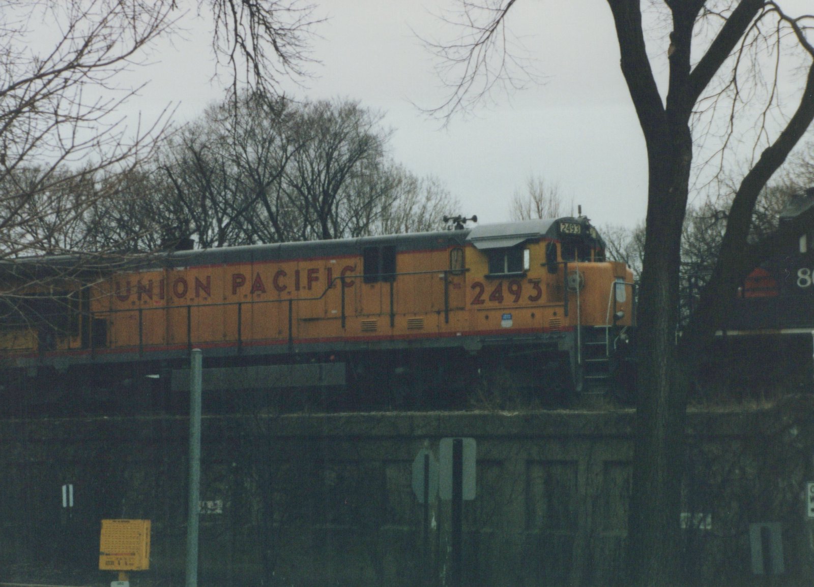 A Union Pacific locomotive, number 2493, painted in yellow, red, and black livery, is traveling on an elevated concrete structure with bare trees in the background.