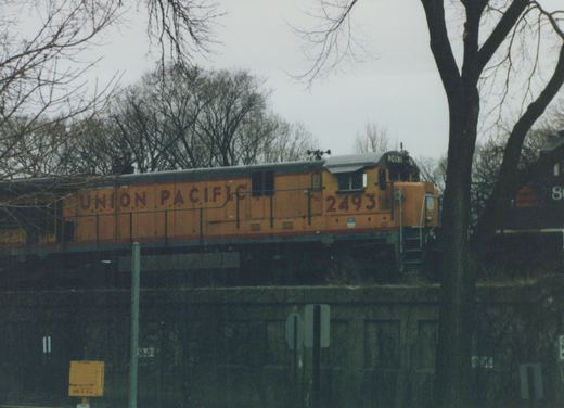 A Union Pacific locomotive, number 2493, painted in yellow, red, and black livery, is traveling on an elevated concrete structure with bare trees in the background.