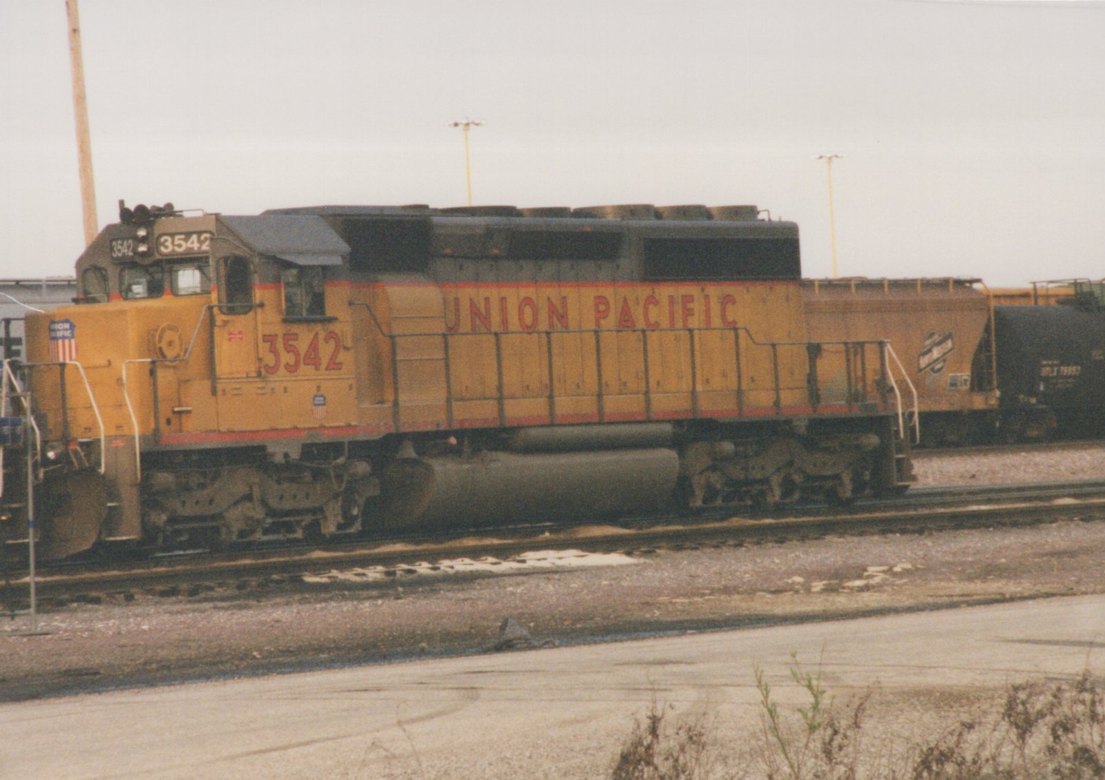 Side view of a yellow Union Pacific locomotive number 3542 on railroad tracks in a yard, with freight cars behind it under an overcast sky.