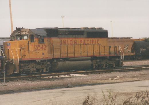 Side view of a yellow Union Pacific locomotive number 3542 on railroad tracks in a yard, with freight cars behind it under an overcast sky.