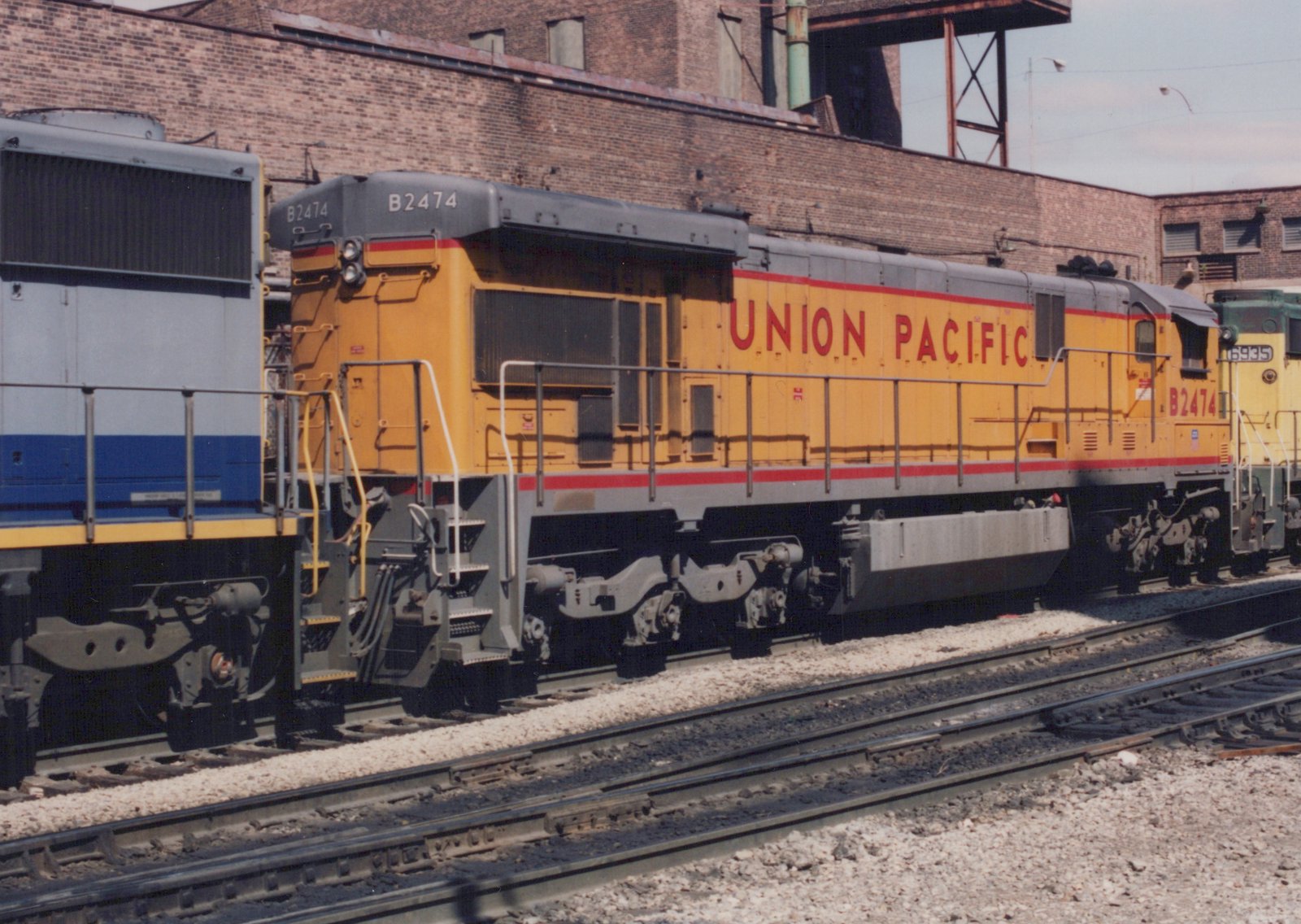 A yellow and gray Union Pacific diesel locomotive numbered B2474 is parked on tracks in a rail yard next to other locomotives, with a brick building in the background.