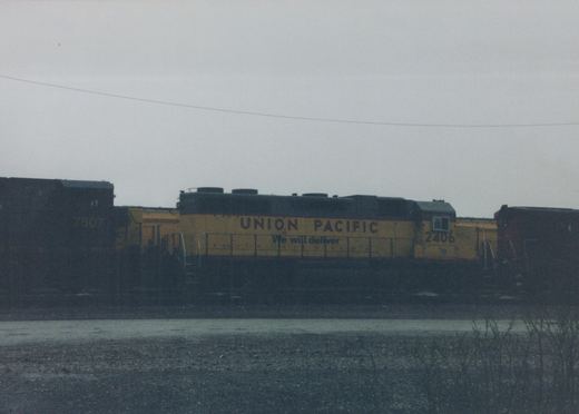 Side view of a yellow and gray Union Pacific diesel locomotive, number 2406, parked on tracks in a yard with another locomotive visible behind it.
