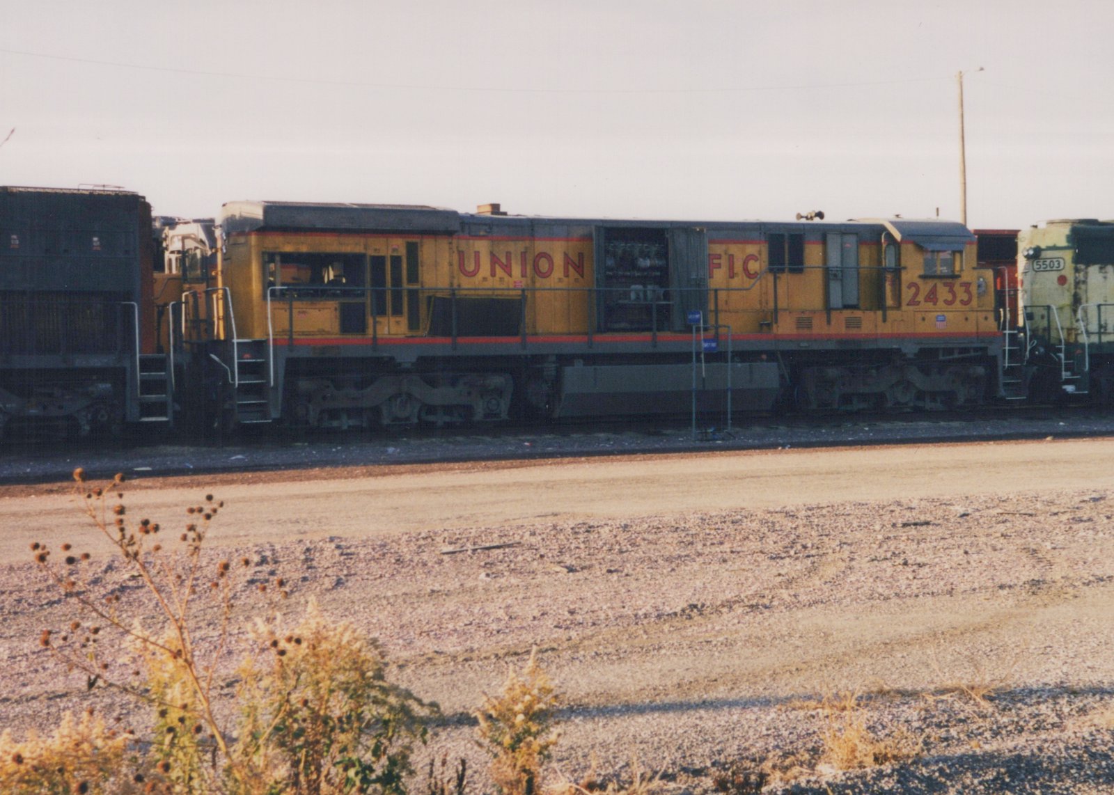 Side view of a yellow and gray Union Pacific diesel locomotive, number 2433, sitting in a gravel rail yard next to another locomotive, number 5503, under a clear sky.