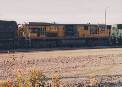 Side view of a yellow and gray Union Pacific diesel locomotive, number 2433, sitting in a gravel rail yard next to another locomotive, number 5503, under a clear sky.