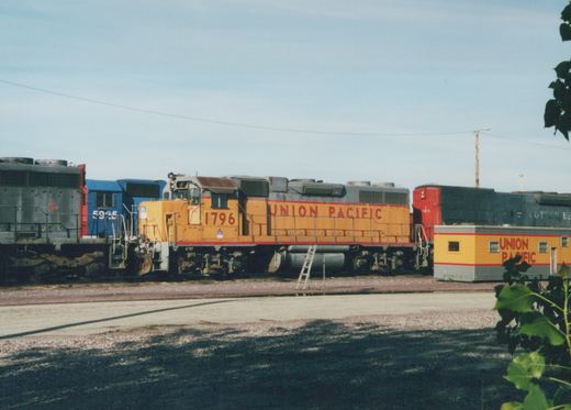 A yellow Union Pacific diesel locomotive with the number 1796 is parked on a track in a rail yard, with other locomotives and a yellow office car visible nearby under a clear sky.