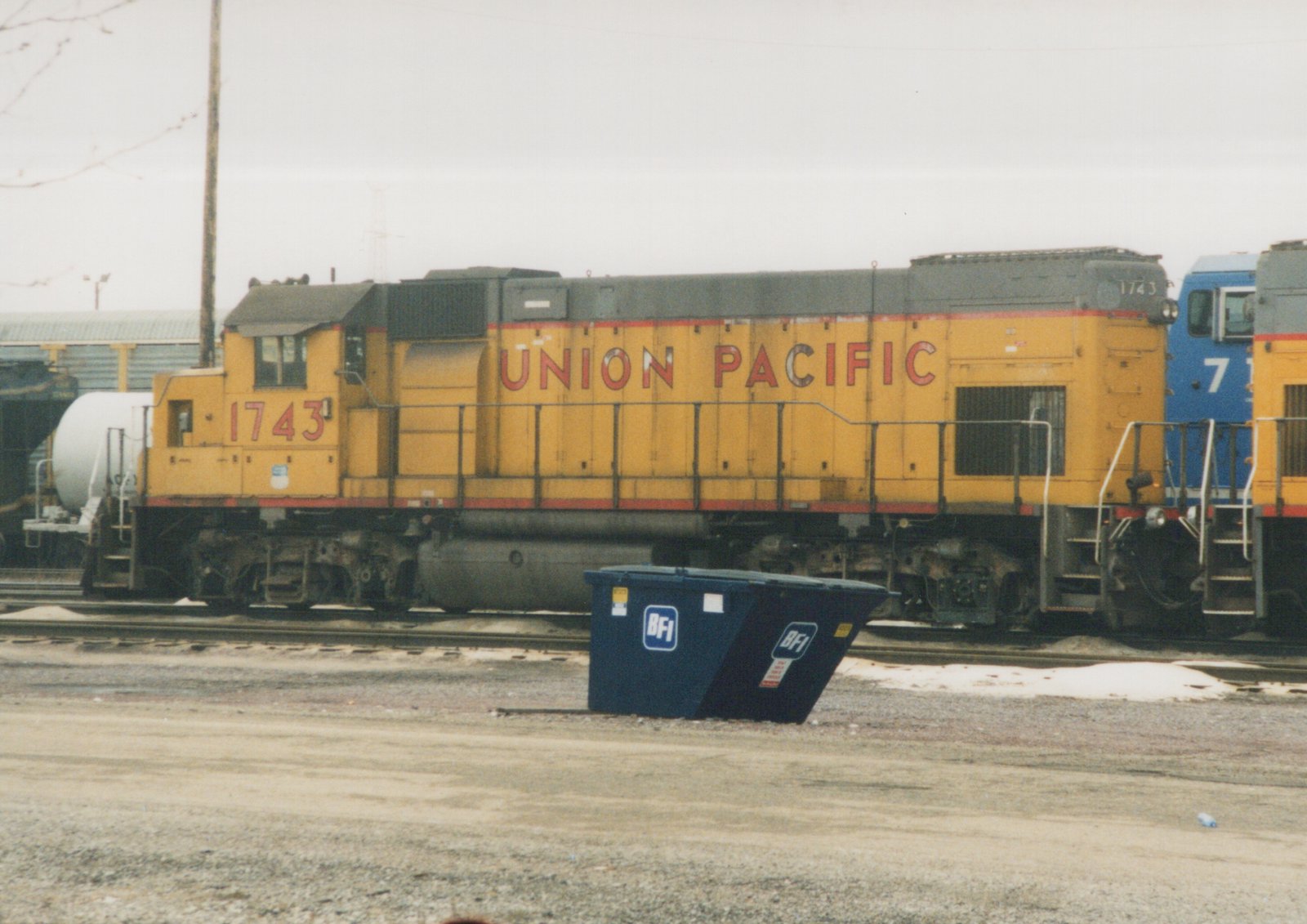 A yellow and gray Union Pacific diesel locomotive numbered 1743 is parked on a railroad track next to a blue dumpster with BFI lettering; a second blue and yellow locomotive is partially visible behind it.
