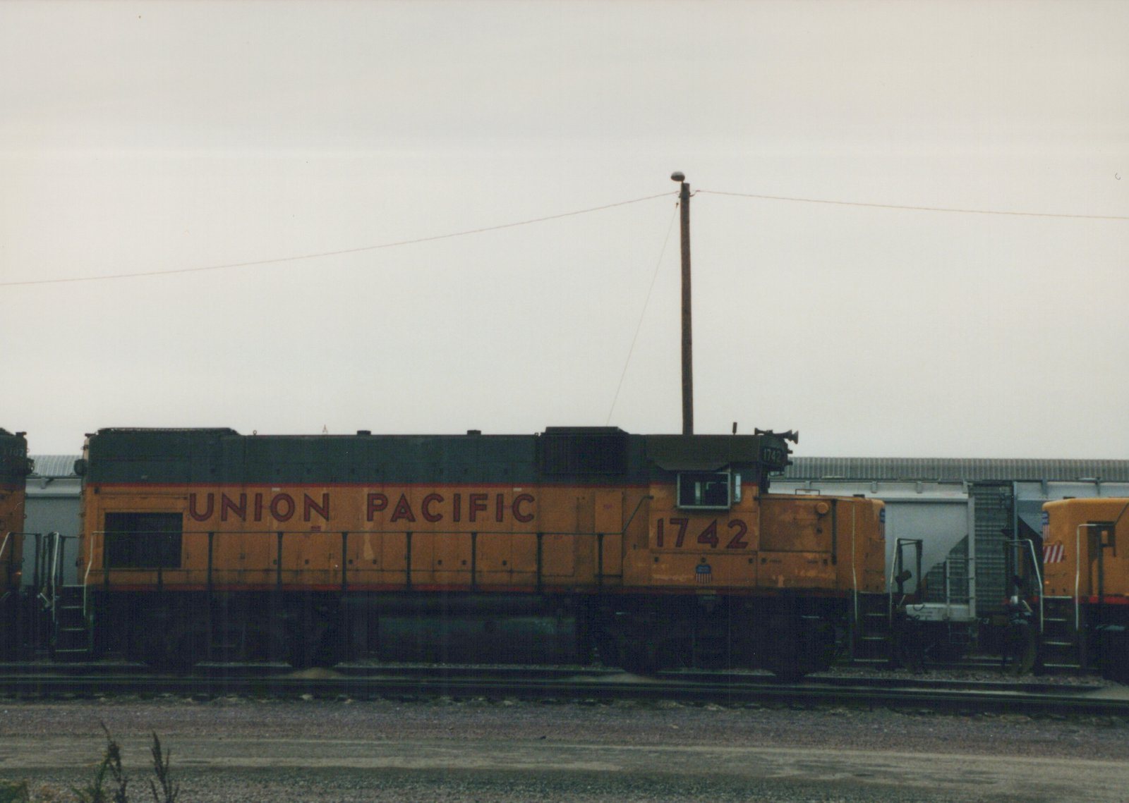 A side view of a Union Pacific locomotive numbered 1742, painted in orange with black and red trim, parked on tracks in a rail yard with a second locomotive and freight car nearby.