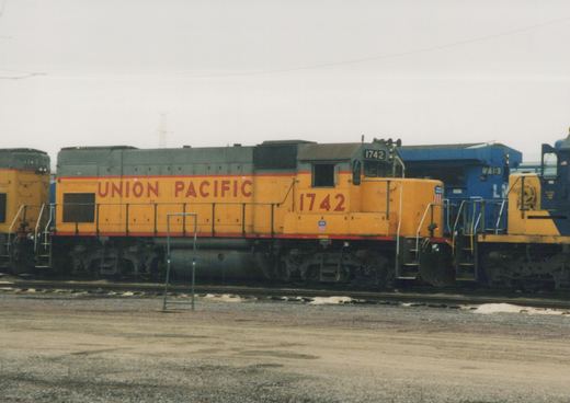 Side view of a yellow Union Pacific diesel locomotive, number 1742, parked in a rail yard with snow patches on the ground and other locomotives nearby.
