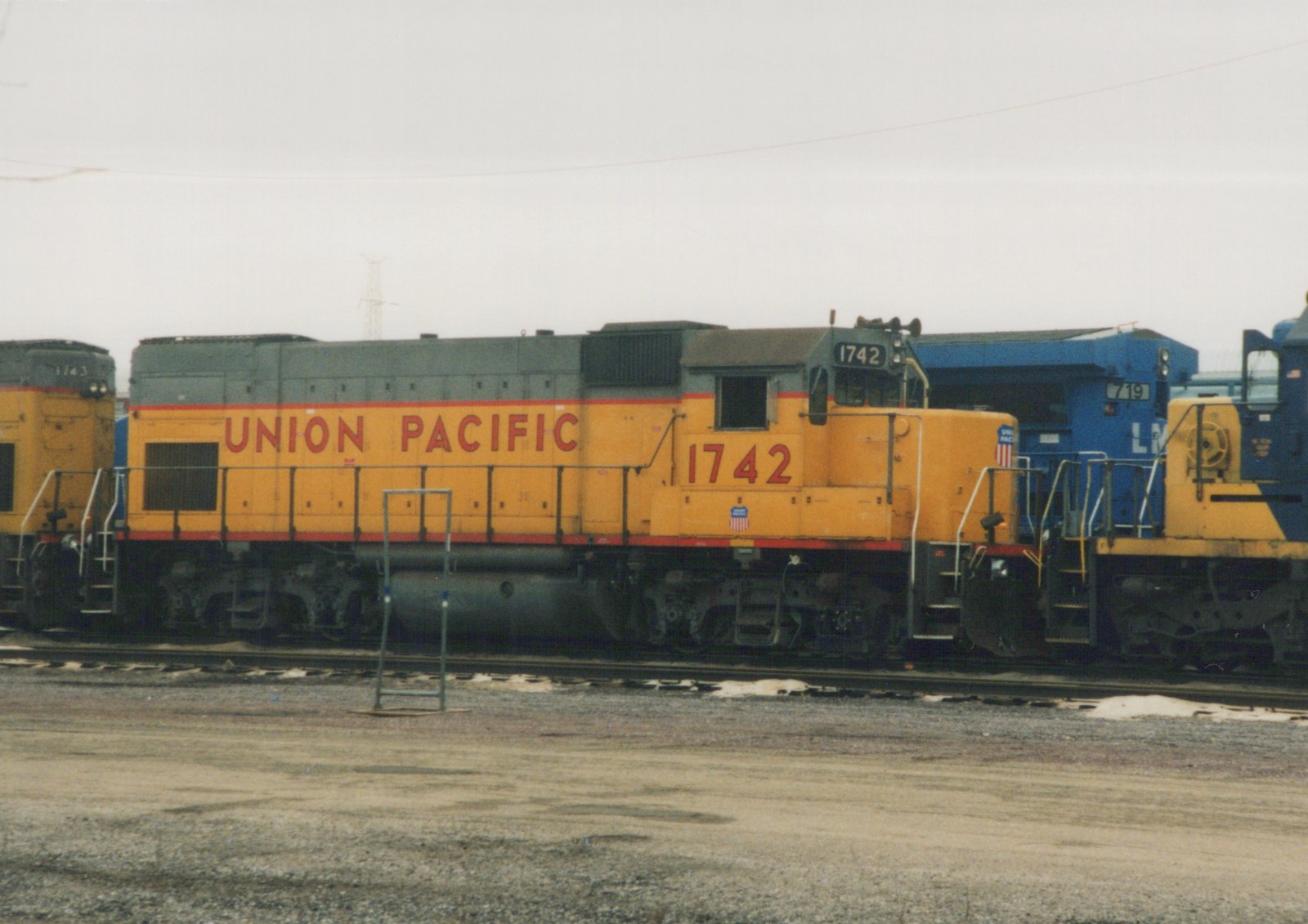 Side view of a yellow Union Pacific diesel locomotive, number 1742, parked in a rail yard with snow patches on the ground and other locomotives nearby.